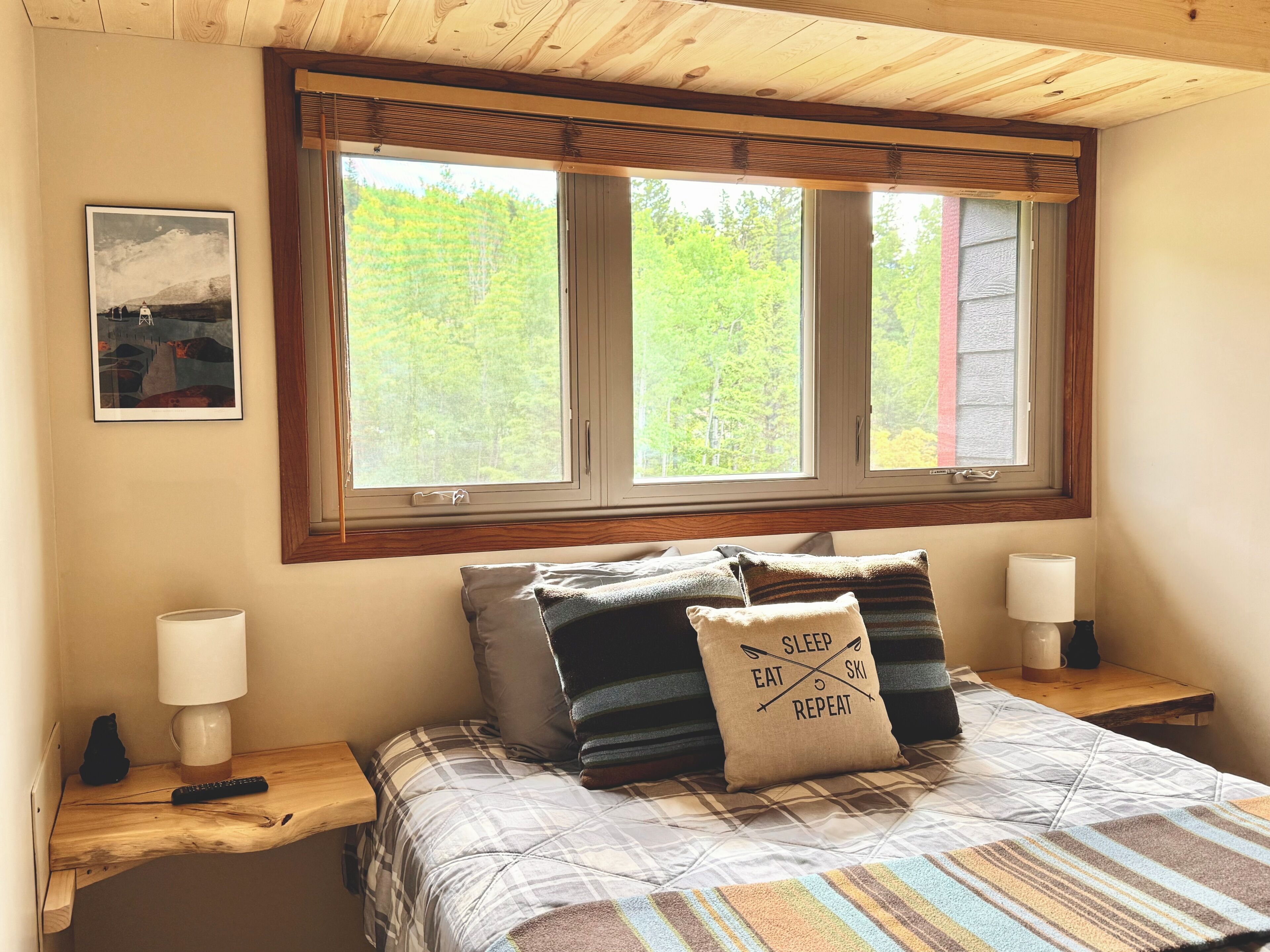 A bedroom with a large window, a bed with plaid bedding, two bedside lamps, and a pillow with text.