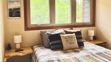 A bedroom with a large window, a bed with plaid bedding, two bedside lamps, and a pillow with text.