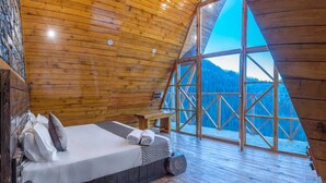 Ein gemütliches Schlafzimmer in einer Holzhütte mit Bett, Kissen und Blick auf einen verschneiten Berg durch große Fenster.