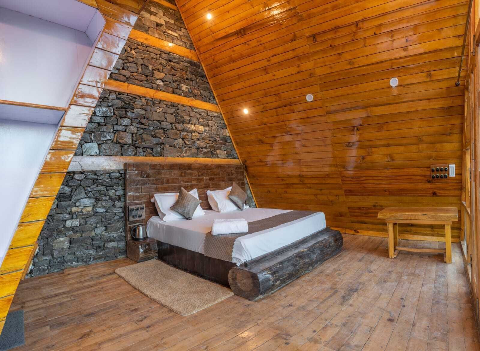 Une pièce triangulaire en bois, chaleureuse et accueillante, avec un mur en pierre, un lit avec des oreillers et un banc en bois.