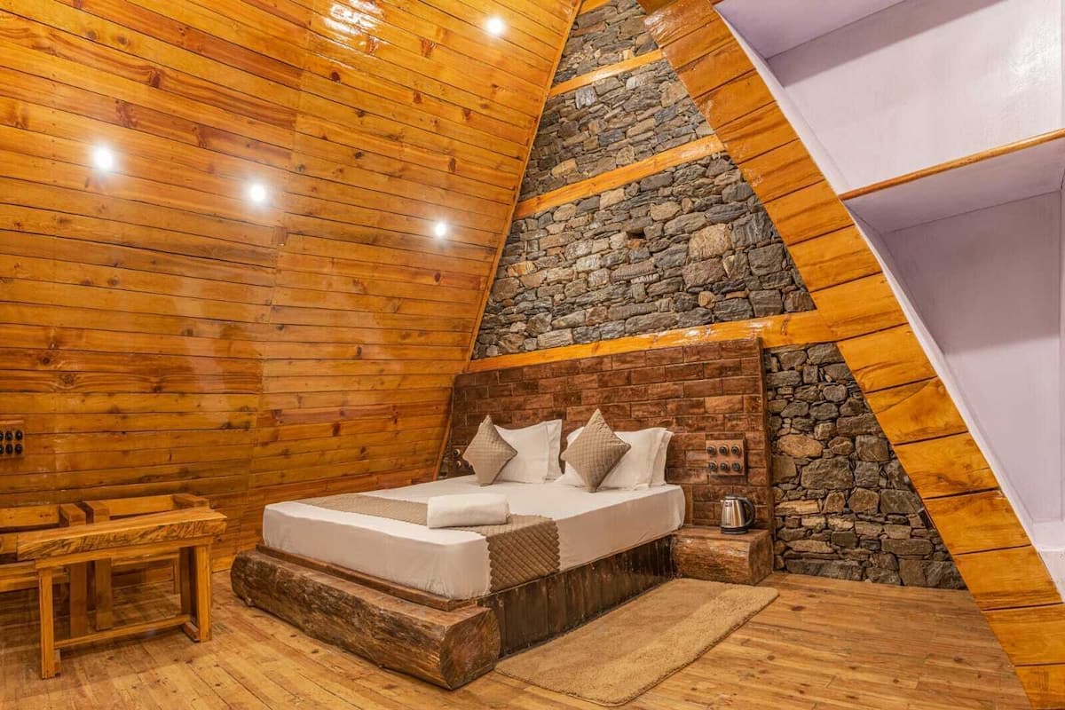A cozy bedroom with a sloped ceiling, wooden walls, a stone accent wall, a bed with pillows, a bedside table, and a nightstand with a kettle.