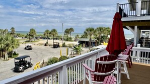 A deck with red Adirondack chairs and a red umbrella overlooking a beach area with palm trees and a golf cart. - Pirate-Themed Luxury Beach House-Free Golf Cart-Ocean Views-Ocean Lakes (Myrtle Beach)