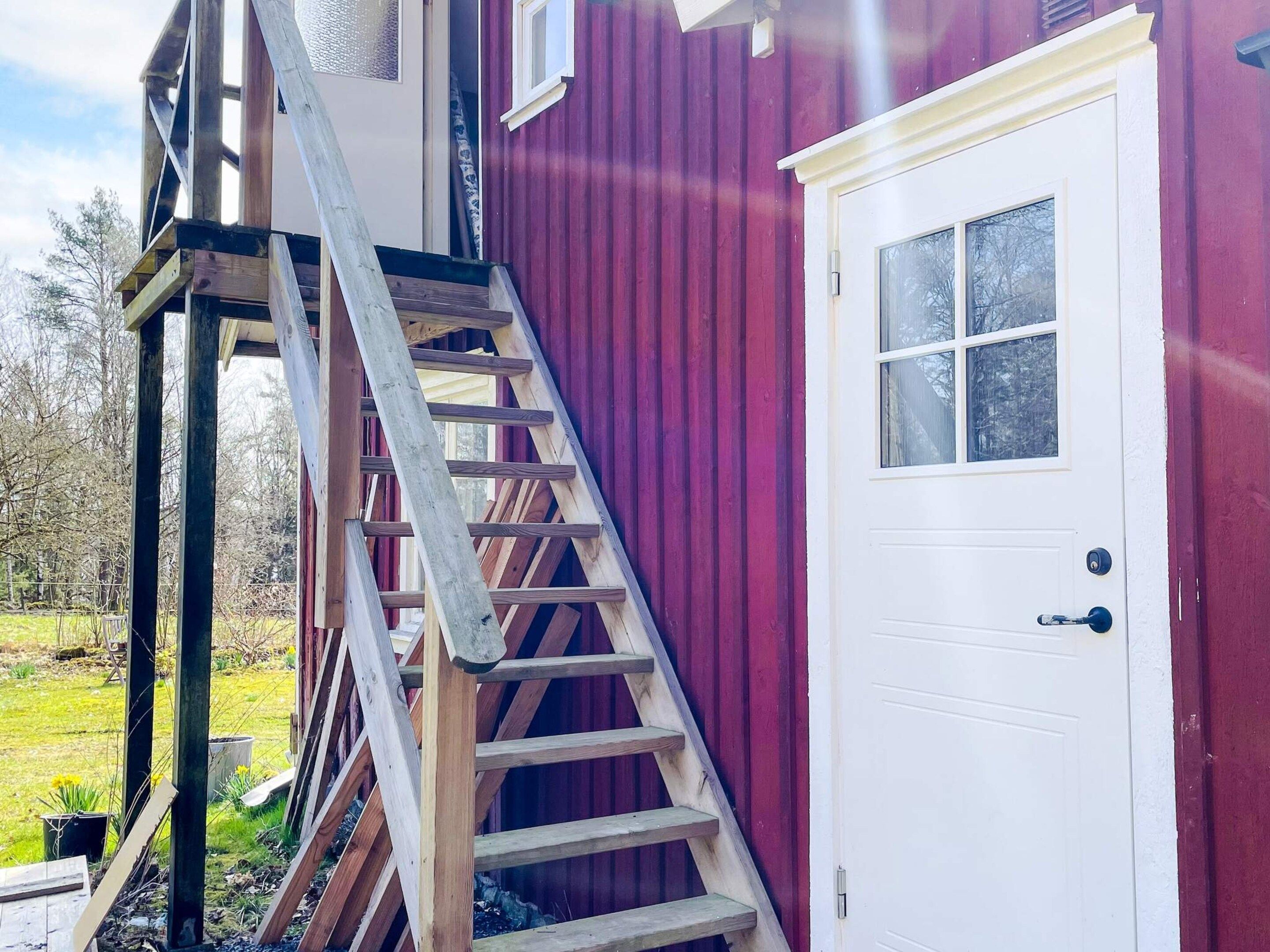 Una escalera de madera que lleva a una puerta blanca con un ventanal, junto a un edificio rojo.