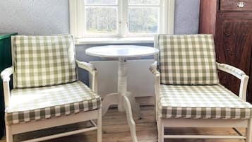 Dos sillas blancas con tapicería verde y blanca a cuadros, una mesa redonda blanca y una ventana con vista a árboles.
