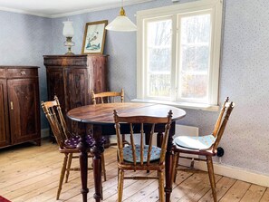Een eetgedeelte met een ronde houten tafel en stoelen, een raam met uitzicht op bomen en een kastje met een foto erboven.