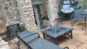 Une terrasse extérieure avec des murs en pierre, une plateforme en bois, une table à plateau de verre et des chaises en métal.
