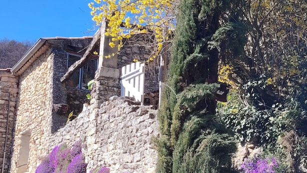 Un bâtiment en pierre avec un portail blanc, entouré de verdure et de fleurs violettes.