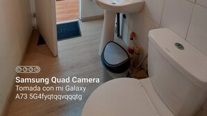 Une salle de bain avec un lavabo, des toilettes et un miroir, tous de couleur blanche. Une porte donne sur un couloir où se trouvent une bibliothèque et une fenêtre.