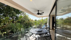 A glass-enclosed balcony with a round table and chairs, a ceiling fan, and a view of a swimming pool and lush greenery. - Modern Condo Jungle Retreat (Puerto Aventuras)