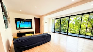 A modern living room with a large window, a flat-screen TV, a sofa, and a small table. - Modern Condo Jungle Retreat (Puerto Aventuras)