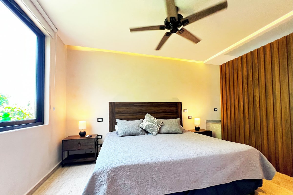 A modern bedroom with a large bed, two bedside tables with lamps, a ceiling fan, and a window with a view of greenery.