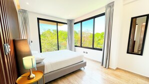 A modern hotel room with a large bed, a bedside table with a lamp, and a view of greenery through large windows. - Modern Condo Jungle Retreat (Puerto Aventuras)
