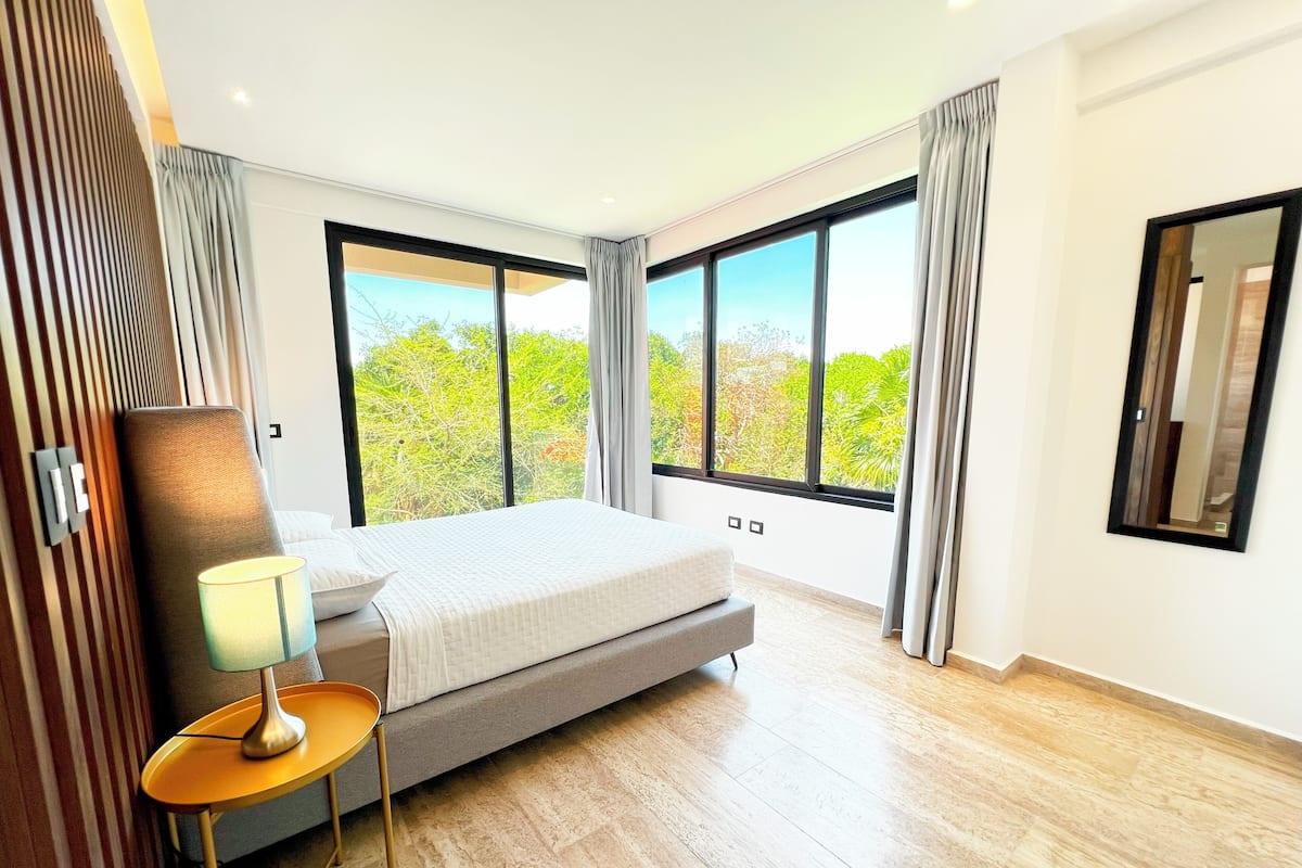 A modern hotel room with a large bed, a bedside table with a lamp, and a view of greenery through large windows.