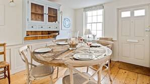 Un comedor con una mesa de madera rústica dispuesta para cuatro personas, rodeada de sillas blancas. Contra la pared, hay un armario blanco con puertas de cristal, y se ve una ventana con cortina blanca.