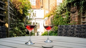 Dos copas de vino sobre una mesa blanca con un cojín estampado, situadas en un jardín con edificios de ladrillo al fondo.