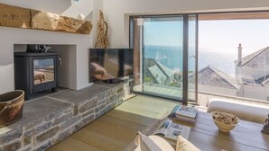 A modern living room with a stone fireplace, a flat-screen TV, and a large window overlooking the sea. - Stylish coastal retreat with panoramic views & walkable to harbour (Mevagissey)