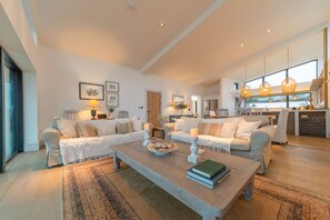 A spacious living room with a large sofa set, a coffee table with books, and a kitchen area in the background.