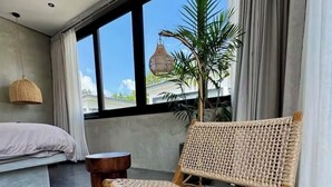 A room with a large window, a wicker chair, a small wooden table, and a woven rug. - 3-bedroom villa in fabulous Uluwatu Bali with pool (Bali)