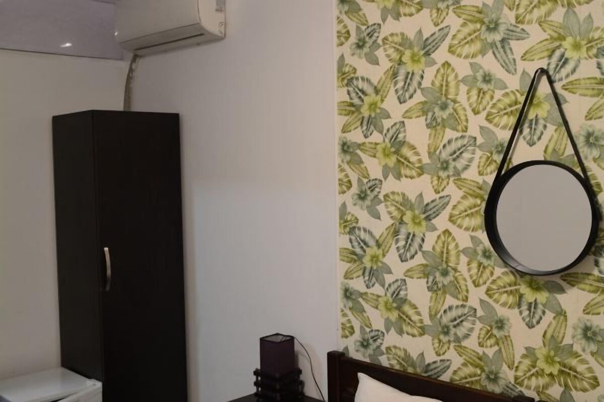 A room with a wardrobe, a bed with a pillow, a wall-mounted round mirror, and a floral wallpaper pattern.