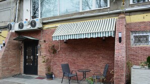 A brick building with a black door, a striped awning, and outdoor seating with chairs and tables. - Hotel Nino on Tsamebuli (Tbilisi)