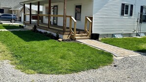 A white single-story house with a covered porch, a flagpole with an American flag, and a gravel driveway. - Quaint Village 4 bedroom home in Friendship IN (Versailles)