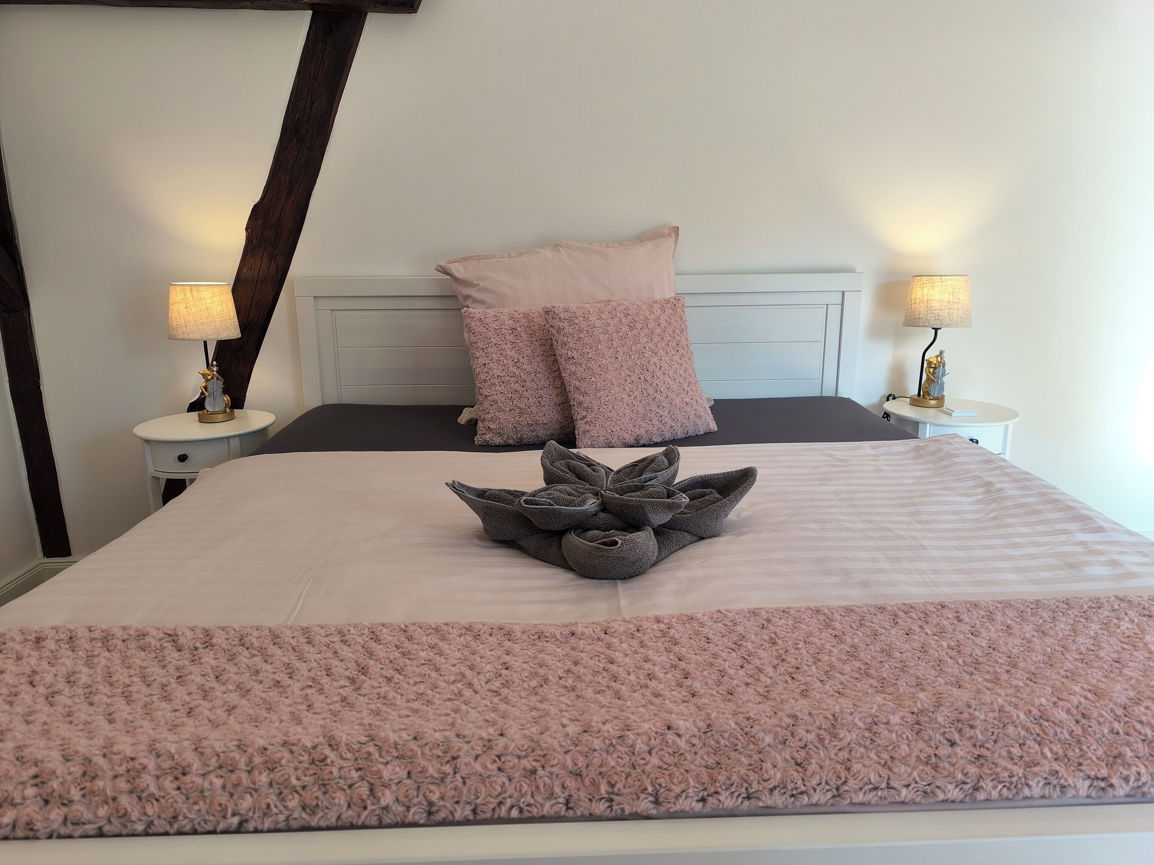Ein ordentlich gemachtes Bett mit einer rosa Decke und einem dekorativen Handtuch in der Mitte, zwei Nachttischlampen und ein Kopfteil mit dunklen Holzapplikationen.