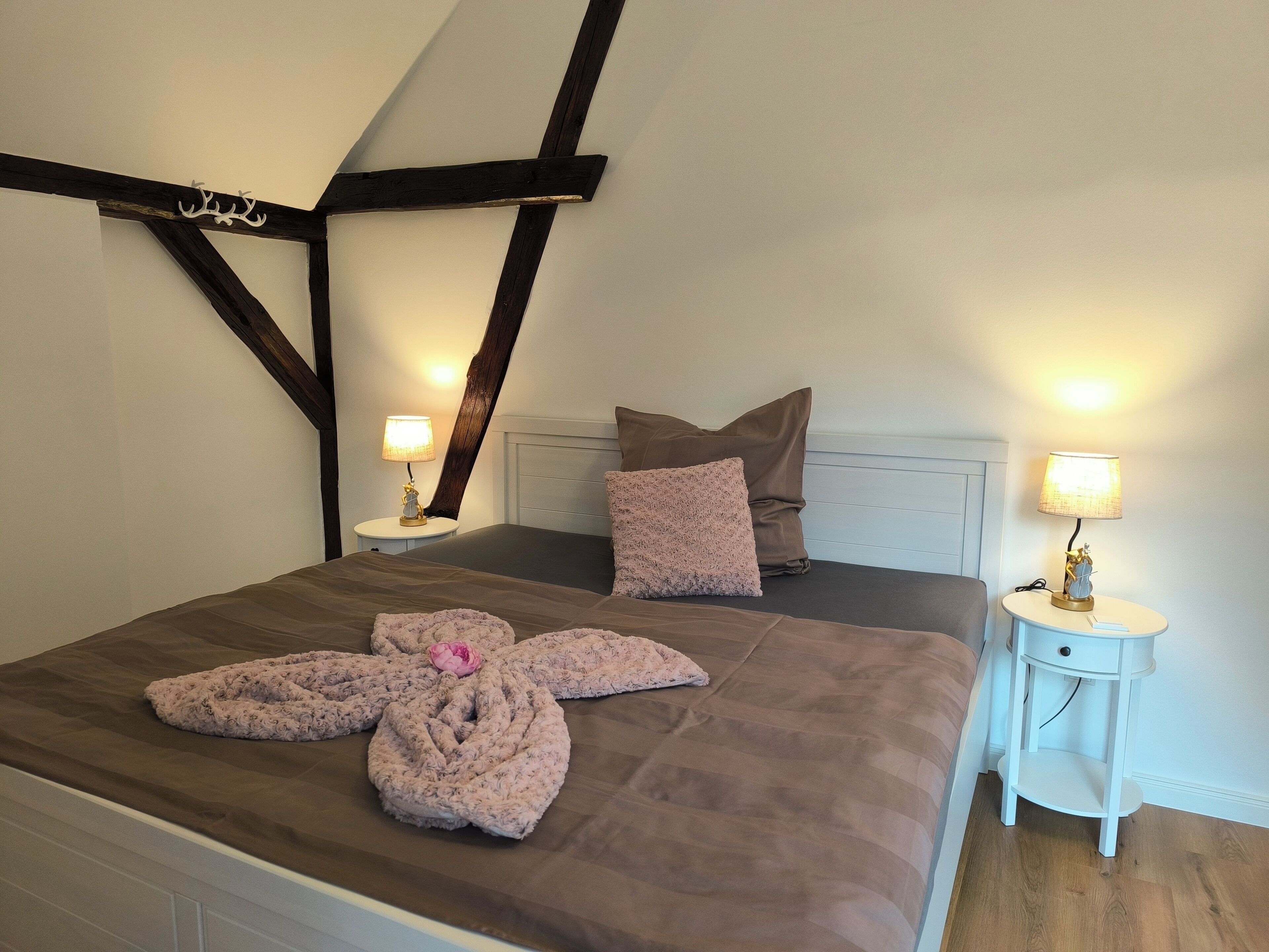 Une chambre avec un plafond en pente, un lit recouvert d’une couverture marron et agrémenté de coussins décoratifs, deux tables de chevet avec des lampes, et une petite table de nuit blanche.