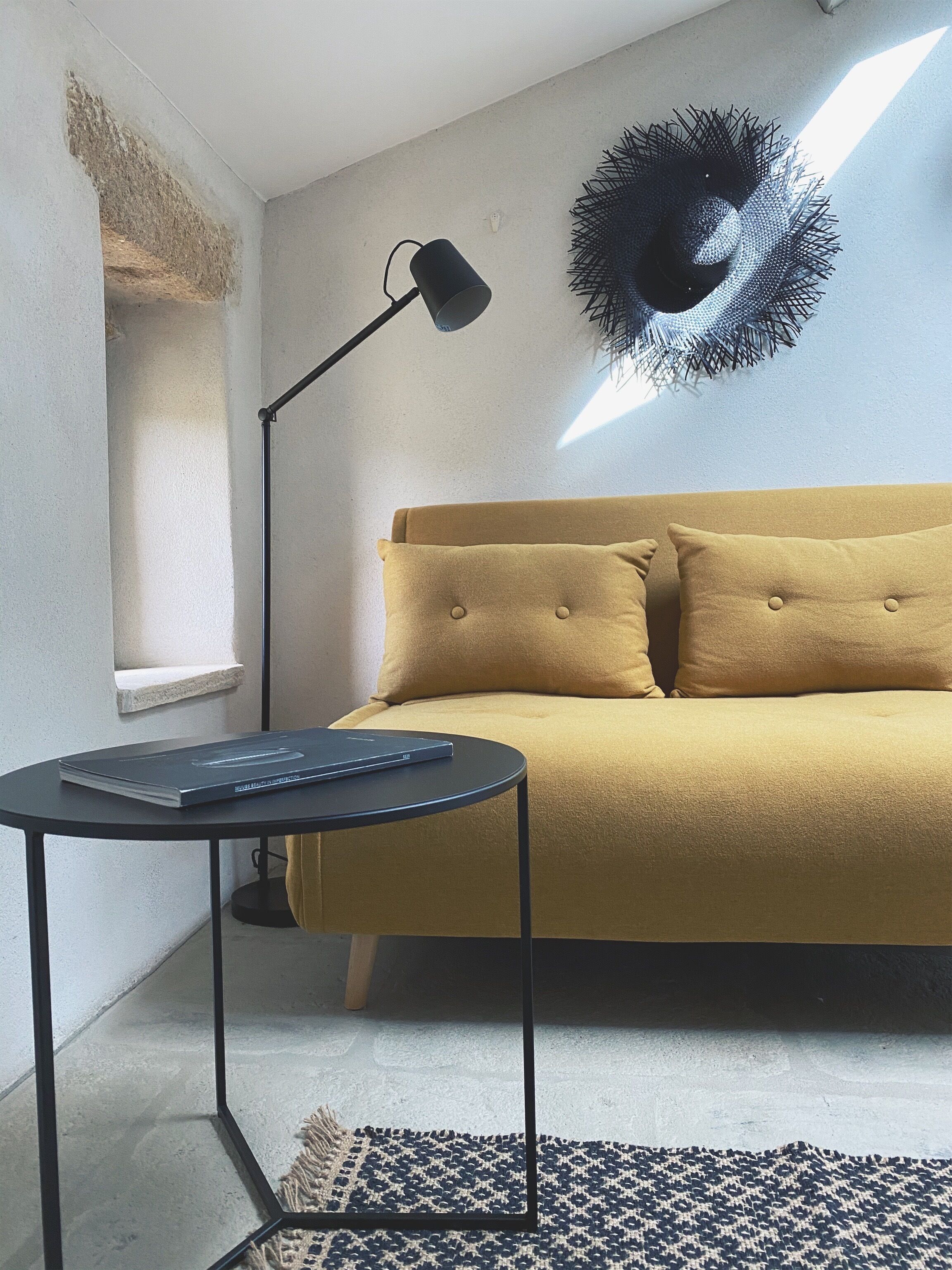 A room with a sofa, a small table with a book, a floor lamp, a wall art piece, and a textured rug.