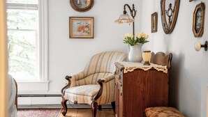 A room with a wooden floor, a striped armchair, a wooden cabinet, a bench, a vase with flowers, and a lamp. - Farmhouse With Heated Pool on Almost 160 Acres! (Elkhorn)