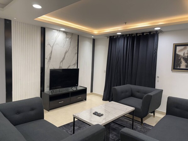 Uma sala de estar moderna com uma TV de tela plana, sofás escuros, uma mesa de centro de mármore e uma janela grande com cortinas.