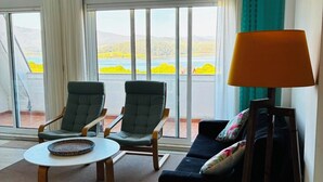 Una habitación con ventilador de techo, un sofá, dos sillones, una lámpara, una mesita y grandes ventanales con vistas a la vegetación y montañas.
