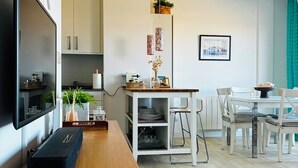 Una sala de estar moderna con televisor, un banco de madera, un comedor con sillas y una mesa, y una cocina al fondo.