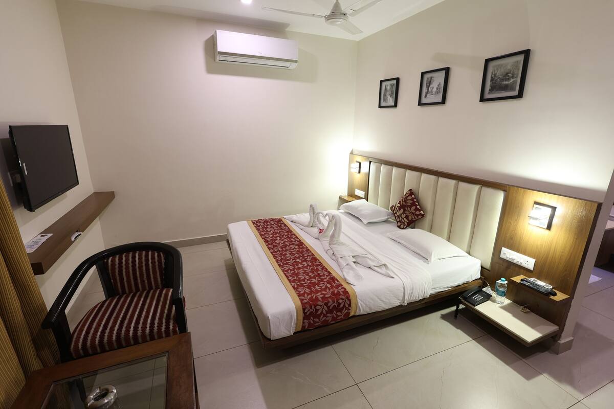 A hotel room with a bed, a chair, a TV, an air conditioner, and a nightstand with a phone and a bottle.