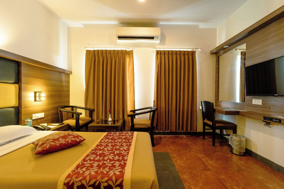 A hotel room with a bed, two chairs, a desk, an air conditioner, and a television.