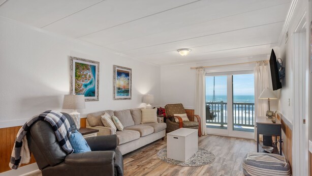 Una sala de estar con sofá, sillón y reposapiés, una mesita y vistas al mar a través de un ventanal.