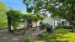 A garden with a paved walkway, a white building, and a tree with green leaves. - Find tranquility and peace in this charming cottage in Lapscheure - Damme. (Damme)