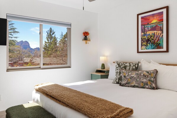 A bedroom with a large window, a bed with white linens, a green throw blanket, and a framed painting on the wall.