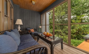 A cozy porch with a wooden ceiling, a cushioned sofa, wicker chairs, a small table with fruit, and a view of lush greenery.