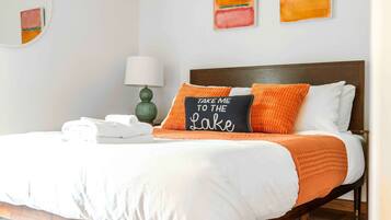 A neatly made bed with white bedding and orange accents, a bedside lamp, and a pillow with the text "TAKE ME TO THE LAKE".