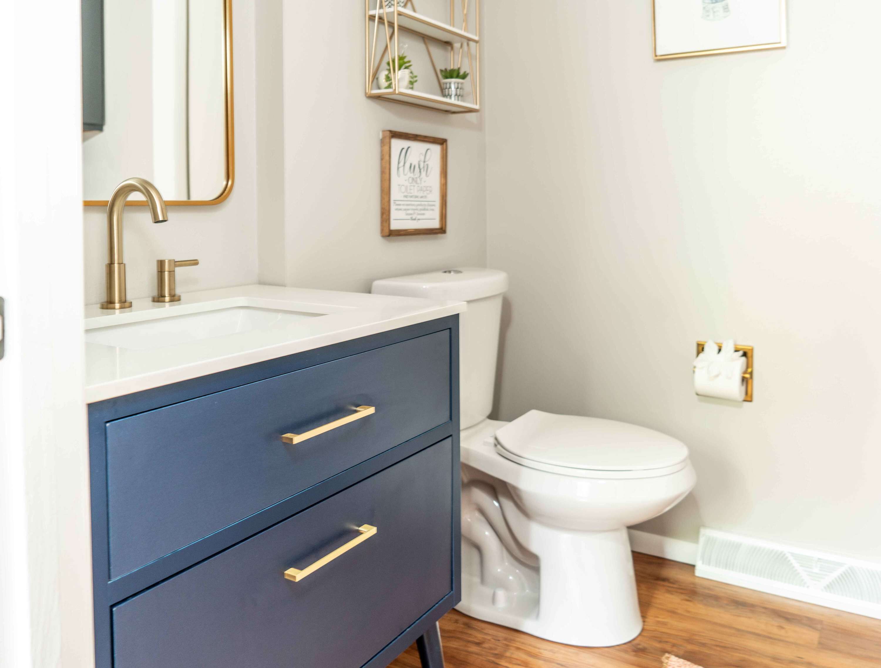 A modern bathroom with a white sink, a dark blue cabinet with gold handles, a white toilet, and a framed sign on the wall.