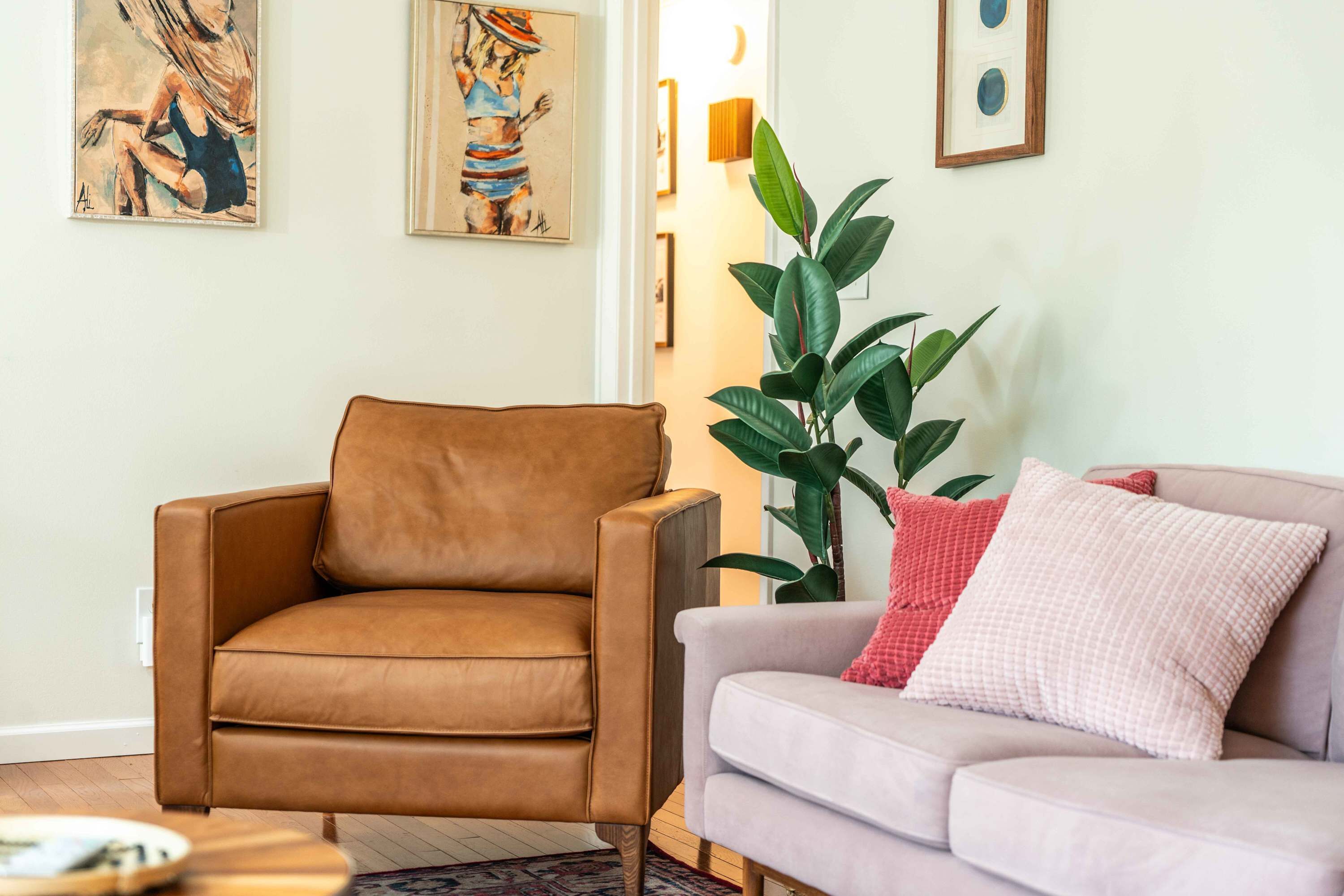 A living room with a brown leather armchair and a light grey sofa with a red pillow. There are two framed artworks on the wall and a large green plant beside the sofa.