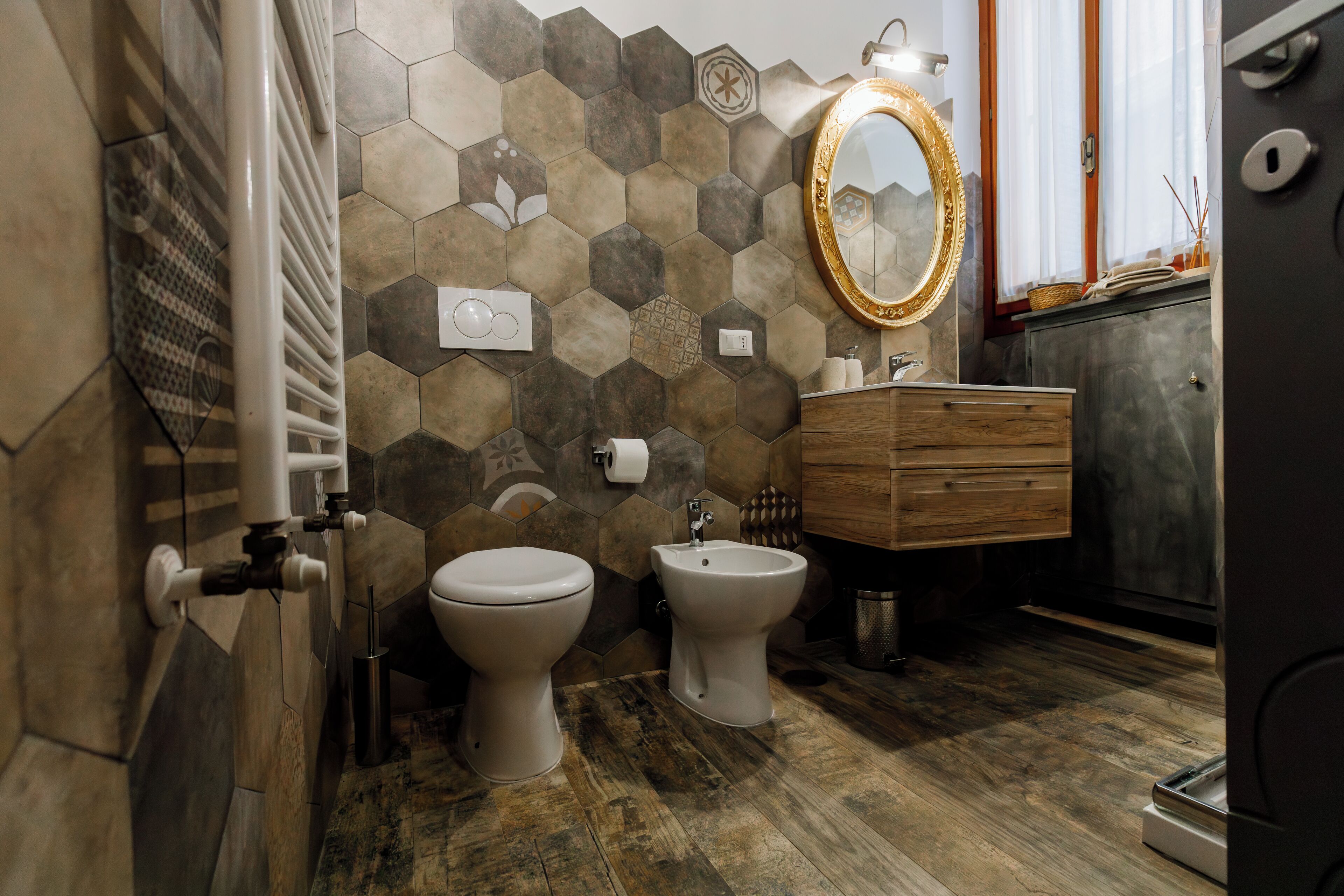 A bathroom with a wooden floor, a white toilet, a bidet, a wooden vanity, and a mirror with a gold frame.