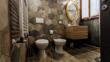 Un baño con suelo de madera, un inodoro blanco, un bidé, un tocador de madera y un espejo con marco dorado.