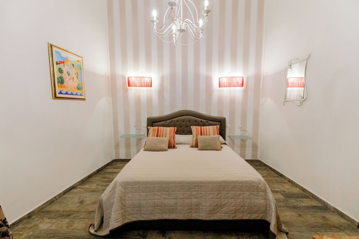 A neatly arranged bedroom with a large bed, two bedside lamps, a chandelier, and a framed picture on the wall.