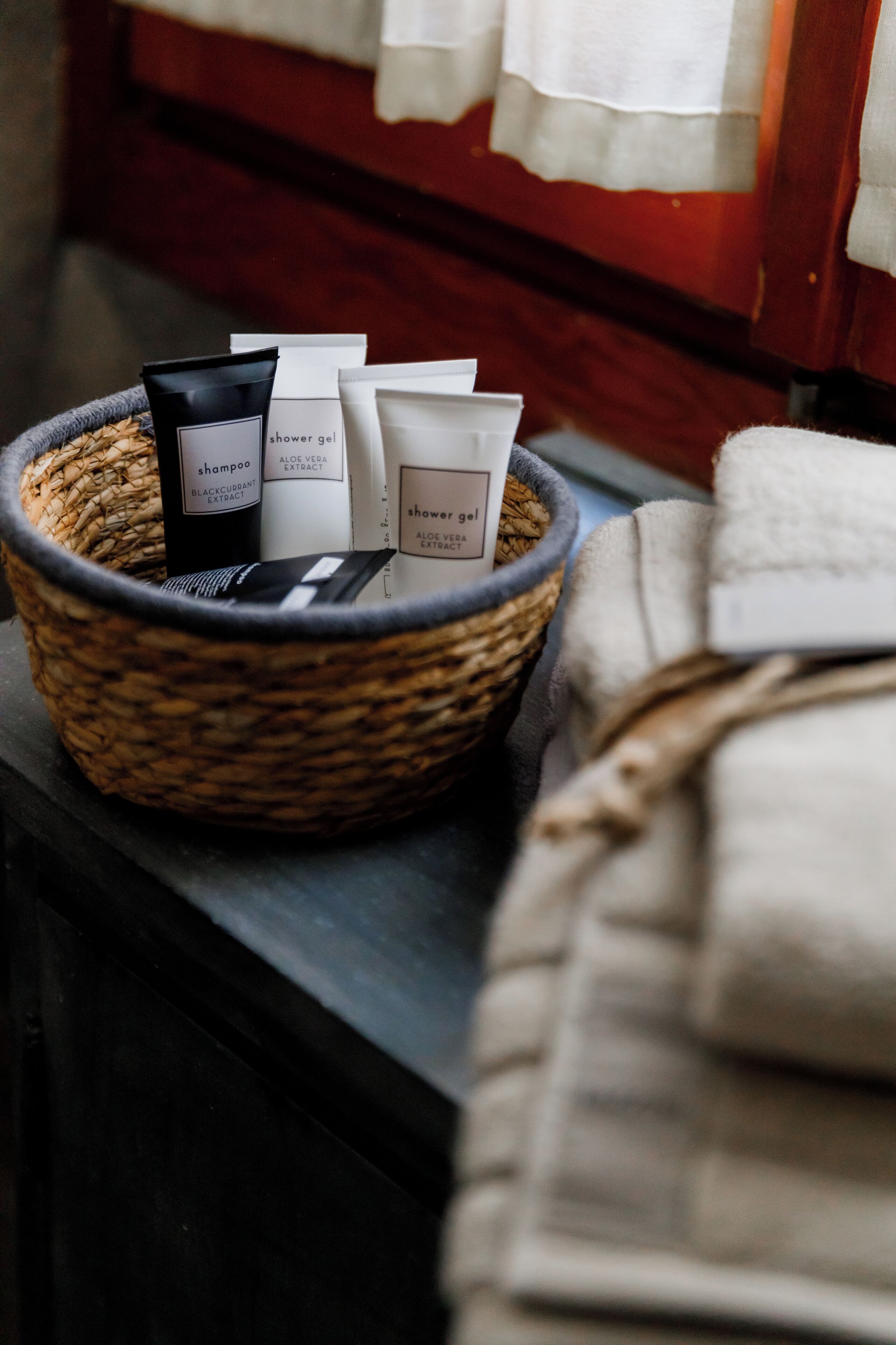 A wicker basket containing toiletries on a dark surface next to a bed with a white blanket.