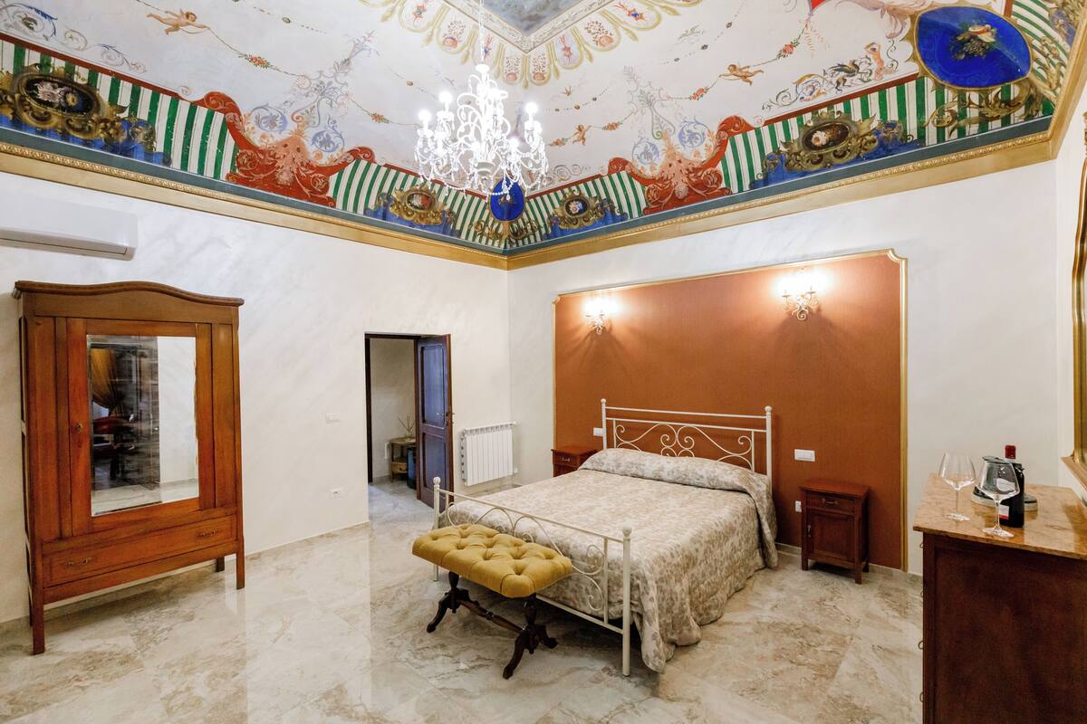 A bedroom with a bed, a wooden wardrobe, a mirror, a chandelier, and a decorative ceiling.