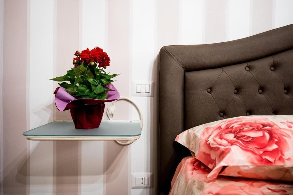A bedroom with a tufted headboard, a bedside table with a glass shelf holding a red flower pot, and a bed with a floral bedspread.