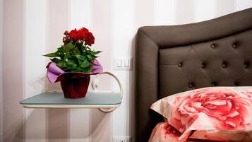 A bedroom with a tufted headboard, a bedside table with a glass shelf holding a red flower pot, and a bed with a floral bedspread.