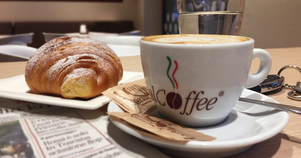A croissant and a coffee cup with a logo on a table with newspapers and a spoon.