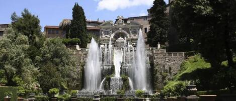 A historic building with a fountain in front, surrounded by lush greenery and a calm water body.
